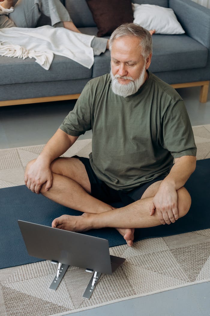 Older man practicing yoga while following an online session on a laptop in his living room.