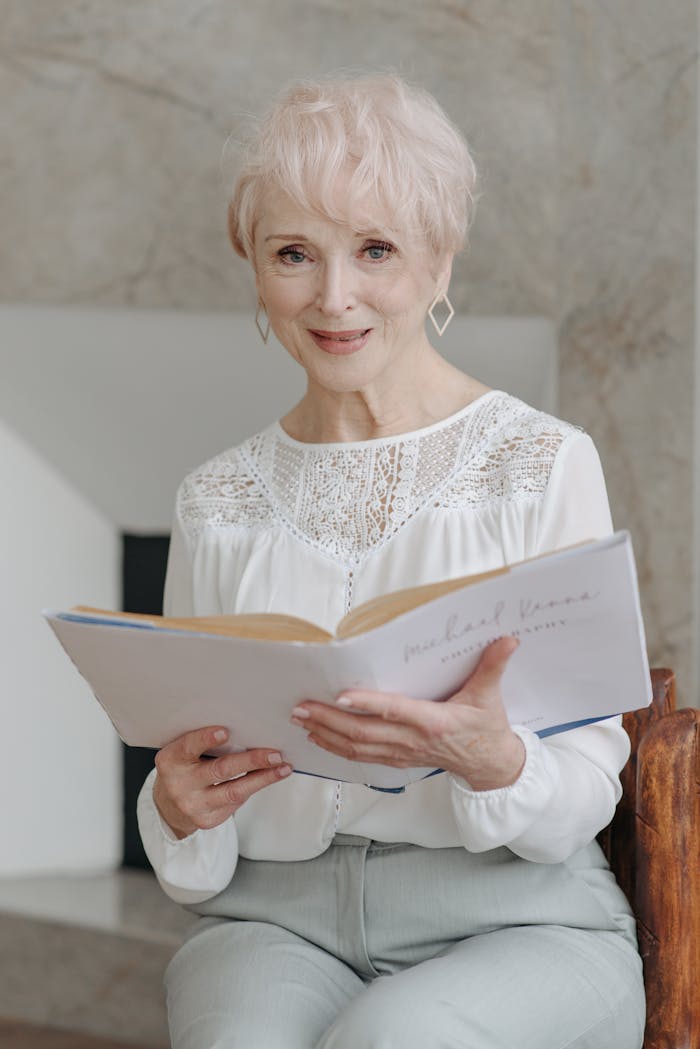 Senior adult woman with white hair reading a book indoors, smiling warmly at the camera.